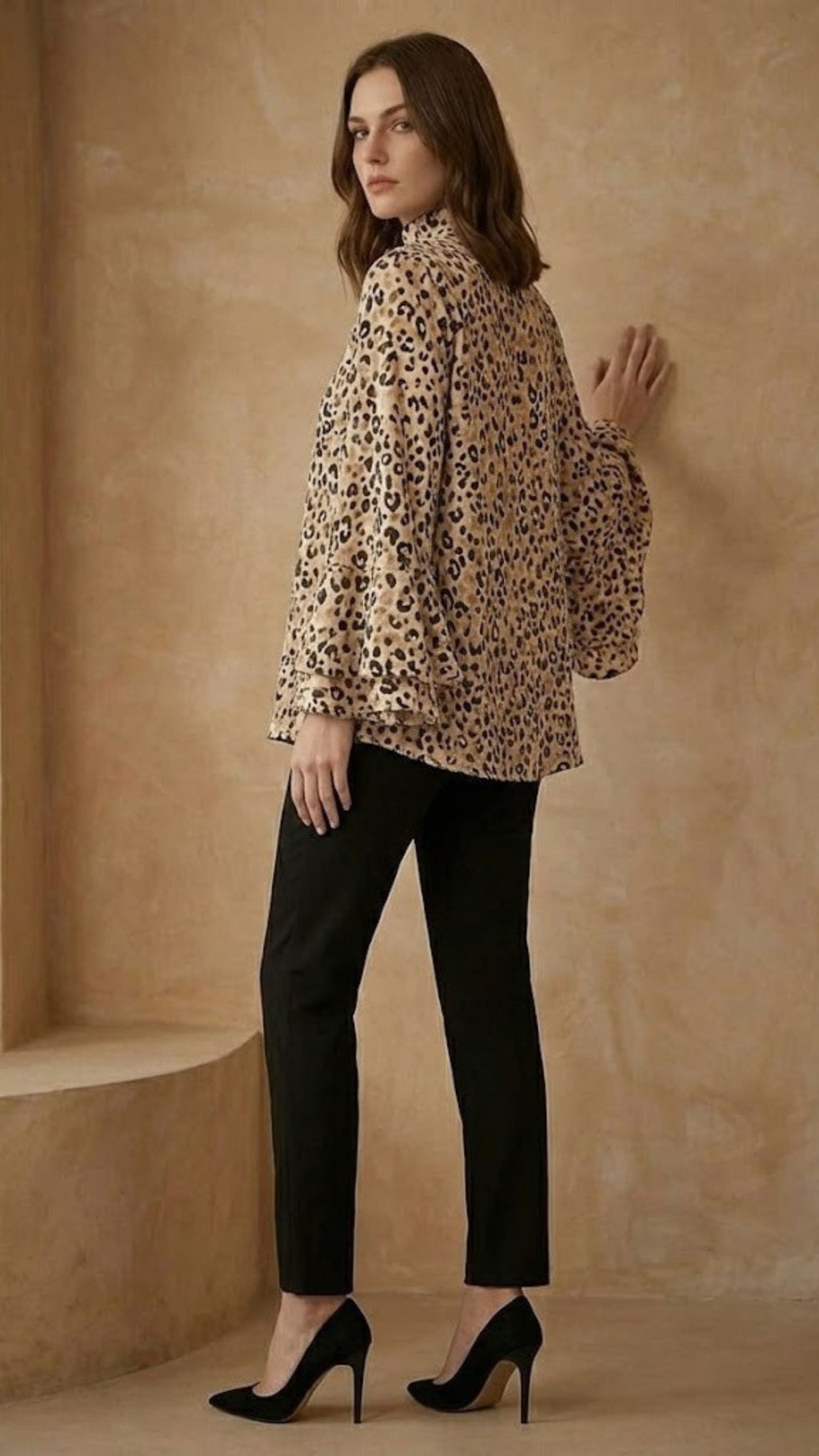 Woman wearing a leopard print blouse and black pants against a beige wall.
