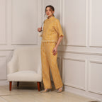 Woman in a yellow patterned outfit standing in a room with white paneled walls and a beige chair.