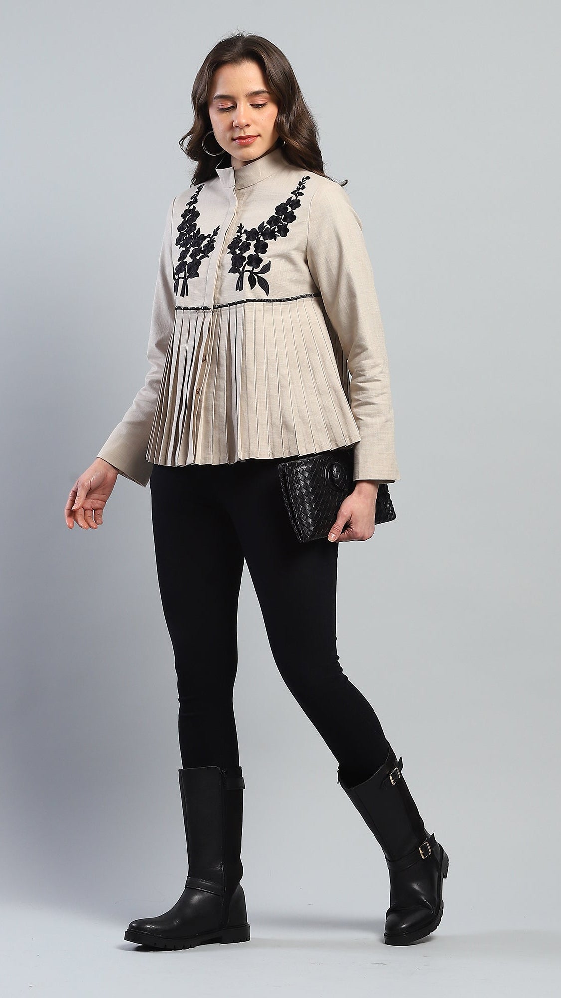 Woman wearing a beige sweater with floral embroidery, black leggings, and black boots on a gray background