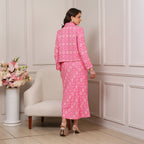 Woman in a pink patterned outfit standing in a room with a white armchair and floral arrangement.