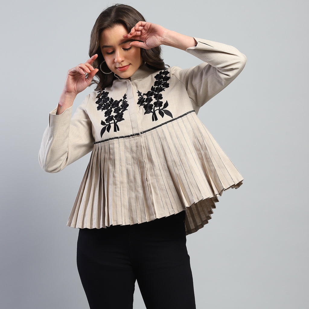 Woman wearing a beige pleated blouse with black floral embroidery on a gray background